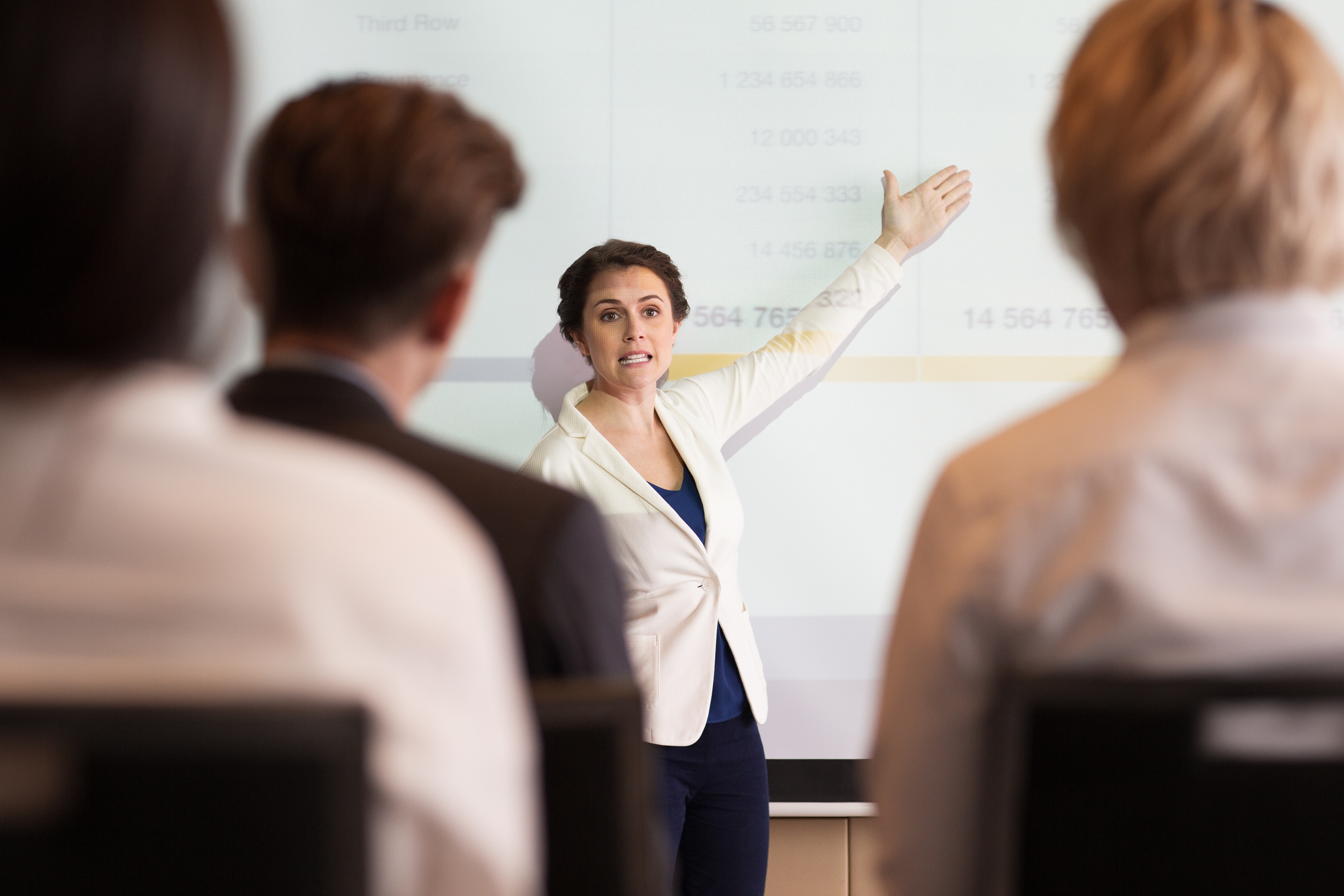 Serious middle-aged businesswoman showing table on projection screen while explaining ideas to audience seen partly